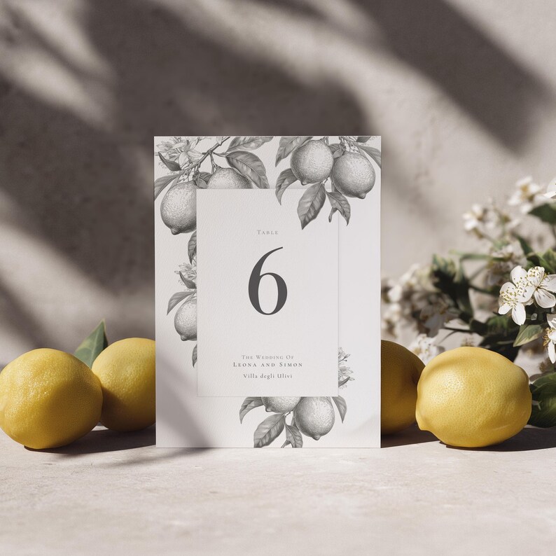 Elegant botanical citrus table number with lemon design, showing the number 6. Propped up in a setting with 4 lemons and lemon blossom.