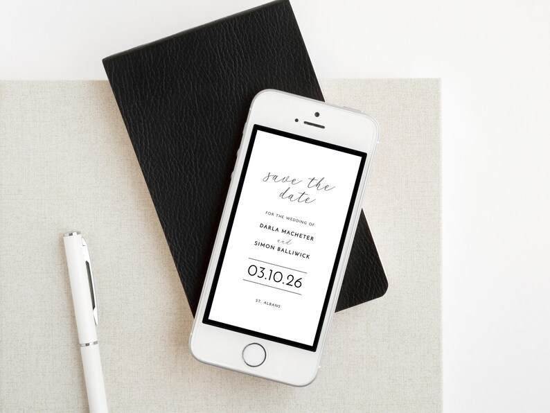 Black border wedding announcement save the date evite displayed on a smartphone. Phone is positioned on top of a black notebook and next to a pen