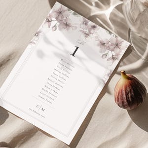 Puede incluir: Una tarjeta de mesa de boda con detalles florales y el número 1. La tarjeta presenta una lista de nombres y la fecha. Un higo maduro y una copa de vino también están en la imagen, sobre una superficie neutra.