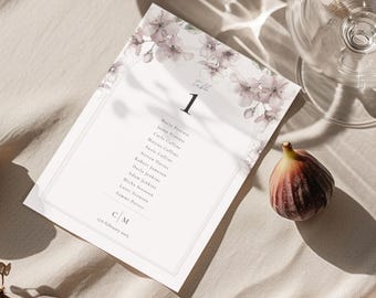 May include: A wedding table card with floral detailing and the number 1. The card includes a list of names and the date. A ripe fig and a wine glass are also in the image, set on a neutral-toned surface.