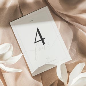 Op de afbeelding: Een witte tafelnummerkaart met het zwarte nummer 4 en het woord "four" in sierlijke letters. De kaart ligt op een beige stof, met witte bloemen op de voorgrond.