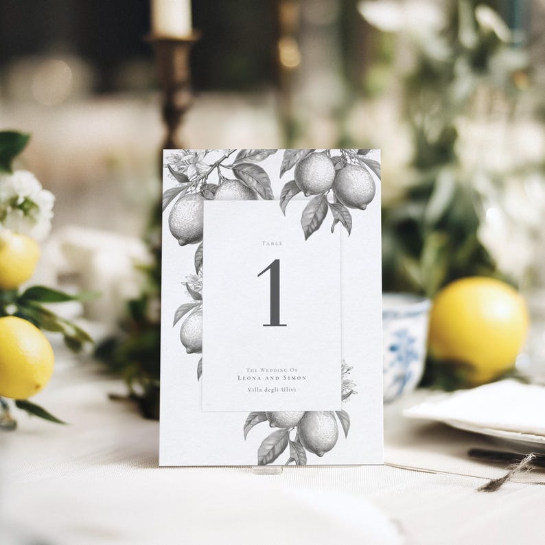 Italian lemon wedding table number in monochrome, showing the number 1 and the names of the happy couple. Shown propped up at a dinner table setting with lemon adornments and candlestick behind.