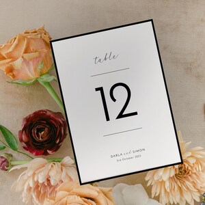 Op de afbeelding: Een witte tafelnummerkaart met een zwarte rand, met het nummer 12 in een vet lettertype. De kaart bevat het woord "table" in een scriptlettertype en de namen Darla & Simon met de datum. De kaart is omgeven door bloemen.