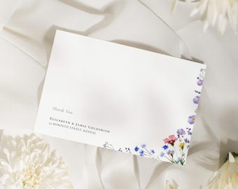 May include: A white card with the words "Thank You" and names and address printed in black. The card is decorated with a floral design in shades of purple, pink, and yellow. The card is on a white fabric surface.