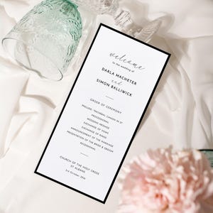 May include: A wedding program with a black border, featuring the names Darla Macheter and Simon Balliwick. The program includes the order of ceremony, with a welcome message at the top. The background is white fabric with a glass and pink flowers.