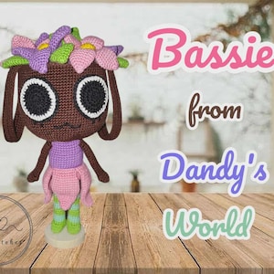 Op de afbeelding: Handgemaakte gehaakte pop genaamd Bassie van Dandy's World. De pop heeft een bruin hoofd met grote zwarte ogen, een paarse top en een roze rok. Het hoofd van de pop is versierd met een bloemenkrans. De tekst "Bassie from Dandy's World" is ook zichtbaar.