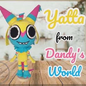 May include: A handmade crochet doll in yellow, blue, and pink, with large black eyes and the text "Yatta from Dandy's World." The doll has a whimsical design with striped horns and a ruffled collar, standing on a wooden surface.