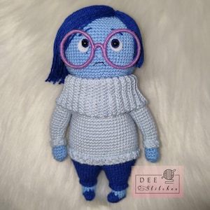 Può includere: Una bambola di uncinetto blu con un'espressione triste, che indossa occhiali rosa e un maglione blu chiaro. La bambola ha pantaloni blu e scarpe blu. La bambola è seduta su una superficie bianca e soffice.