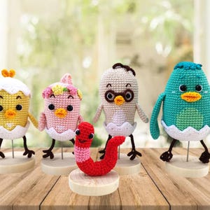 May include: Five crocheted amigurumi toys, including four chicks and a worm, are displayed on wooden bases. The chicks are yellow, pink, white, and blue, and each has a different expression. The worm is red and has a smiling face.