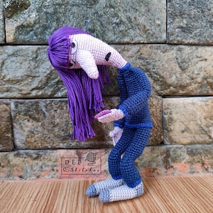 May include: A crocheted doll with purple hair, a pink face, and a blue outfit. The doll is standing with its head tilted down and its arms outstretched. The doll is standing on a wooden surface in front of a stone wall.