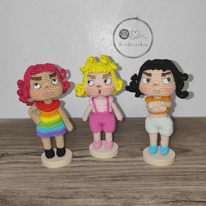 May include: Three handmade crochet dolls with distinctive expressions. One doll is in a rainbow dress, another in a pink outfit, and the third in a white top and shorts. Each doll is on a wooden base. The image includes the text "DeeStitches".