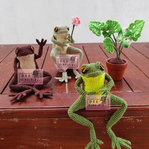 May include: Three crocheted frog figurines in brown, green, and light green, each with orange eyes. One frog holds a pink flower, and another has its arms raised. A crocheted plant in a small pot is also present. The frogs have the text "DEE Stitches" on a tag.