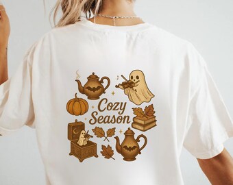 Może przedstawiać: Kremowa koszulka z jesienną grafiką. Projekt zawiera dynię, imbryki, ducha grającego na skrzypcach i napis "Cozy Season". Ilustracje są w odcieniach brązu i beżu.