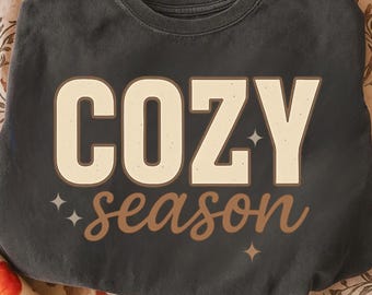 May include: Dark gray sweatshirt with the words "COZY season" in a cream and brown color scheme. The word "COZY" is in a bold, block letter font, and "season" is in a cursive font. Small star graphics are on either side of the words.