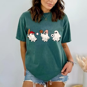 May include: A teal blue t-shirt featuring a graphic of three ghosts playing sports. One ghost is playing baseball, one is playing basketball, and one is playing football.