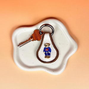 May include: A white and brown embroidered keychain with a bear design, a silver key ring, and a brown key. The keychain is on a white, irregularly shaped dish, set against an orange background.