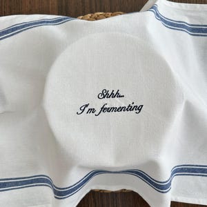 May include: A white kitchen towel with blue stripes draped over a round object. The fabric features black embroidered text that reads, Shhhh... I'm fermenting, centred on the top surface. The towel has a woven texture and clean edges.