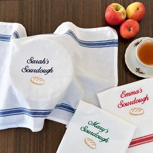 May include: Three white cotton kitchen towels with striped borders in blue, red, and green. Each towel features embroidered text reading Sarah's Sourdough, Emma's Sourdough, or Mary's Sourdough, accompanied by a small embroidered loaf of bread icon.