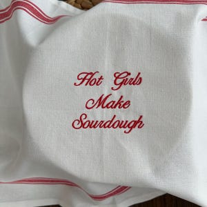 May include: A white kitchen towel featuring red striped borders and the red embroidered text Hot Girls Make Sourdough in a cursive script font. The fabric is draped over a round object, creating soft folds across the surface.