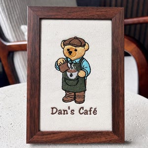 May include: A rectangular wooden frame containing a fabric panel with an embroidered tan bear wearing a brown cap, a blue shirt, and a green apron. The bear is pouring liquid into a white cup. The text Dan's Café is embroidered below the bear.