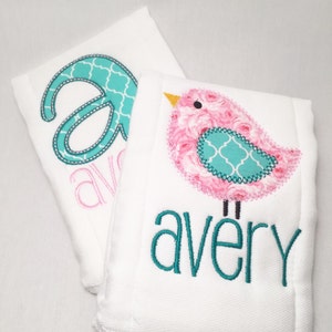 May include: Two white baby burp cloths. One cloth features a large teal letter "A" with a white patterned design, and the name "avery" in pink. The other cloth has a pink bird with a teal patterned design and the name "avery" in teal.