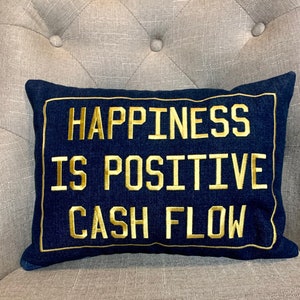 May include: A dark blue denim pillow with a gold embroidered message that reads "Happiness is positive cash flow".