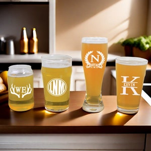 May include: Four personalized beer glasses with different designs. The first glass has a white deer antler design with the letters "WE" inside. The second glass has a white circle with the letters "LNM" inside. The third glass has a white wreath with the letter "N" inside and the text "Best Man 11.06.23". The fourth glass has a white design with the letters "K" and "K" and the word "KEVIN" in between.