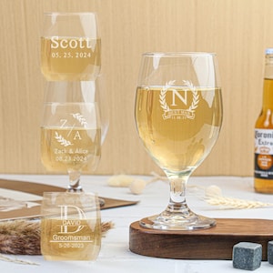 以下が含まれることがあります： 名前と日付が刻まれた、異なるデザインのパーソナライズされたグラスが4つあります。グラスには透明な液体が入っています。ビールのボトルと、灰色の石が置かれた木製のトレイもあります。