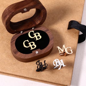 Może przedstawiać: Otwarta, owalna drewniana szkatułka z mankietami z inicjałami w kolorze złotym. Wyświetlane są dwie pary spinek do mankietów z różnymi inicjałami, w kolorze złotym, czarnym i srebrnym. Pudełko ma czarne aksamitne wnętrze.