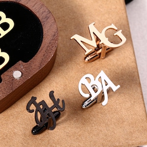Könnte beinhalten: Vier Manschettenknöpfe mit Initialen in Gold, Silber und Schwarz. Die Manschettenknöpfe werden auf einer braunen Oberfläche präsentiert. Ein Paar befindet sich in einer Holzkiste mit goldenen Buchstaben.