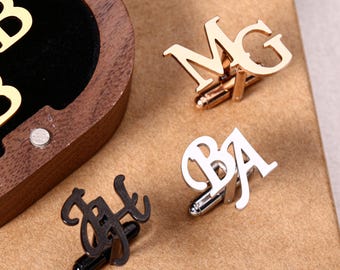 May include: Four pairs of initial cufflinks in gold, silver, and black. The cufflinks are displayed on a brown surface. One pair is in a wooden box with gold letters. The cufflinks are a personalized gift.