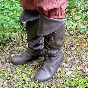 May include: Brown leather knee-high boots with a lace-up design and folded-over tops. The boots are worn with reddish-brown trousers. The boots are on a grassy surface.