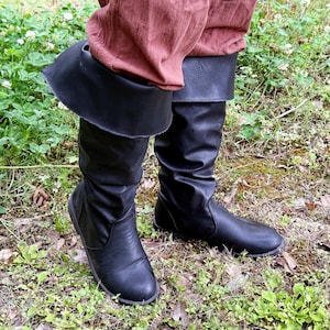 May include: Black knee-high boots with a folded-over cuff. The boots are made of a smooth, synthetic material and are worn with rust-colored pants. The boots are on a grassy surface.