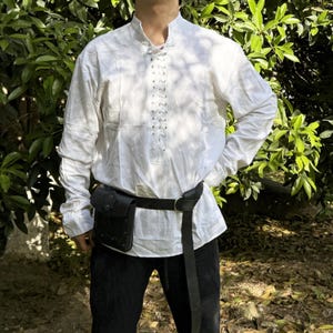 May include: A white long-sleeved shirt with a lace-up front and a black leather belt with a small pouch attached. The shirt has a high collar and is worn by a person standing in a garden setting.