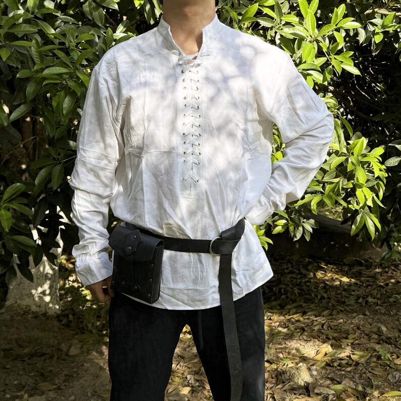 May include: A white long-sleeved shirt with a lace-up front and a black leather belt with a small pouch attached. The shirt has a high collar and is worn by a person standing in a garden setting.
