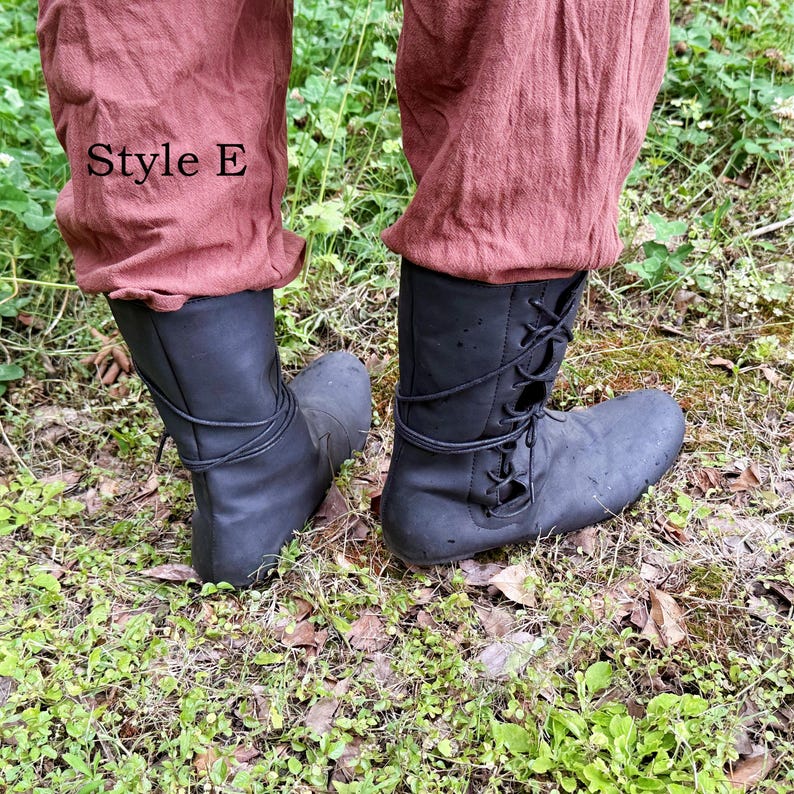 May include: Black leather boots with laces, shown from the ankles down. The boots are worn with rust-coloured trousers. The text "Style E" is visible on the trousers. The boots are on a bed of grass and leaves.
