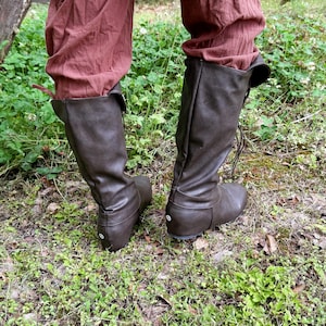 May include: Brown leather knee-high boots with a rounded toe and lace-up closure. The boots are worn with reddish-brown trousers. The boots are set on a bed of green grass and leaves. The boots are a dark brown colour.