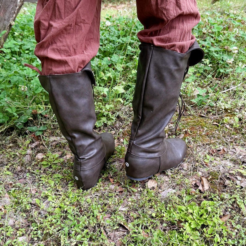 May include: Brown leather knee-high boots with a rounded toe and lace-up closure. The boots are worn with reddish-brown trousers. The boots are set on a bed of green grass and leaves. The boots are a dark brown colour.