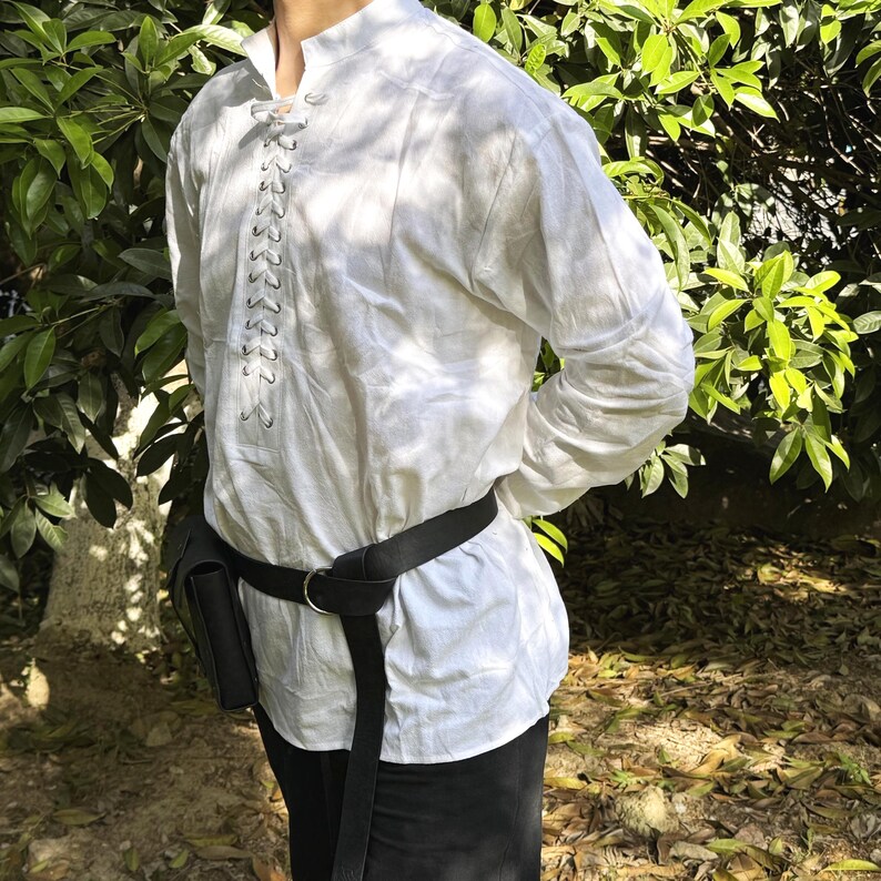 May include: A white long-sleeved shirt with a lace-up detail at the neckline. The shirt is worn with a black belt and a black leather bag.