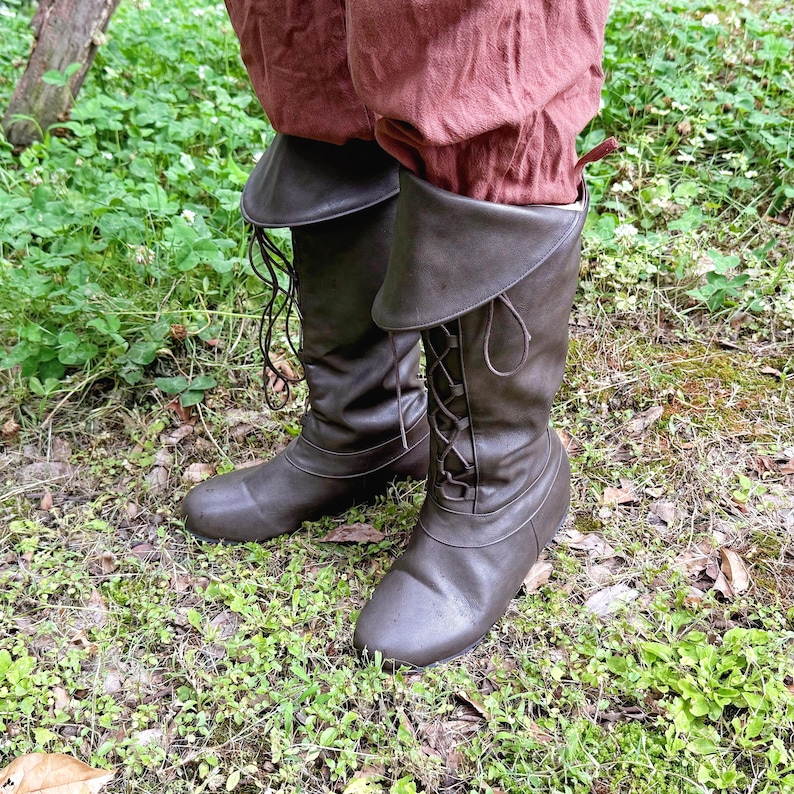 May include: Brown leather knee-high boots with a lace-up design and folded-over tops. The boots are worn with reddish-brown trousers. The boots are on a grassy surface.