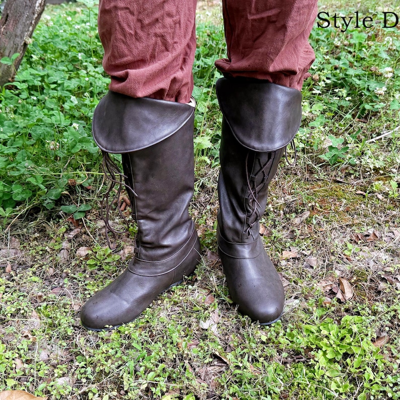 May include: Brown leather knee-high boots with a lace-up design and a folded-over cuff. The boots are worn with burgundy-coloured trousers. The boots are designed in the style of historical footwear.