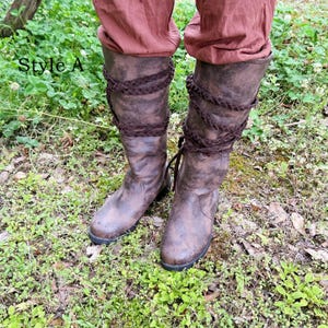 May include: Brown leather knee-high boots with braided detailing. The boots have a distressed appearance and a dark sole. The text "Style A" is visible. The boots are worn over brown trousers and are set against a background of green grass and foliage.
