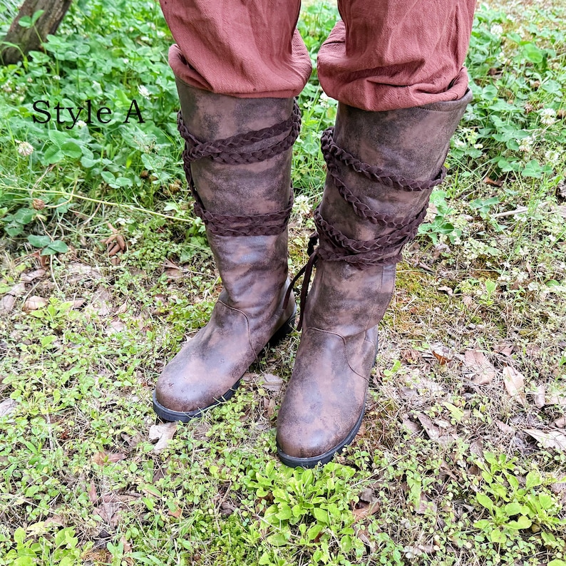 May include: Brown leather knee-high boots with braided detailing. The boots have a distressed appearance and a dark sole. The text "Style A" is visible. The boots are worn over brown trousers and are set against a background of green grass and foliage.