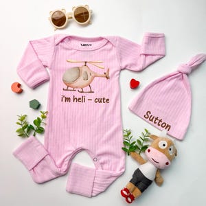 May include: A light pink ribbed long-sleeve romper with a helicopter graphic and the text "i'm heli - cute", a matching pink ribbed beanie with the name "Sutton" printed on it, tan sunglasses, and a small plush cow toy with a white and brown body.