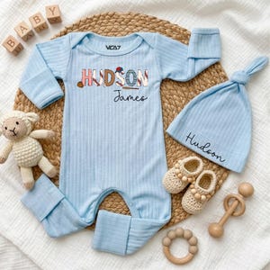 May include: A light blue ribbed long-sleeve baby romper featuring the name Hudson James with baseball-themed graphics, a matching light blue ribbed beanie with the name Hudson, cream-coloured crocheted booties, a small stuffed sheep, and wooden blocks.