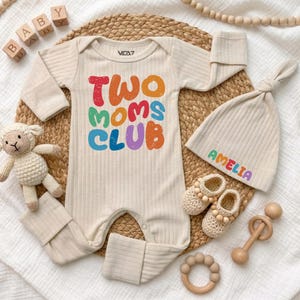 May include: A cream-colored ribbed long-sleeve baby romper with the text TWO MOMS CLUB in colorful letters, a matching ribbed beanie with the name AMELIA, a small crocheted sheep toy, a pair of crocheted booties, and a wooden rattle.