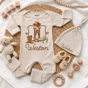 May include: A beige ribbed long-sleeve baby romper featuring a brown graphic of a cowboy hat, boots, cactus, and the letter W with the name Weston. Includes a matching ribbed beanie with the name Weston embroidered, and crochet baby booties.
