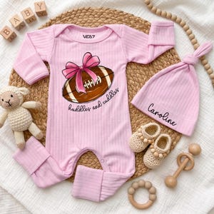 May include: A light pink ribbed long-sleeve romper with a graphic of a brown football and a pink bow, featuring the text 'huddles and cuddles'. Includes a matching pink ribbed beanie with the name 'Caroline' and a pair of beige crochet booties.