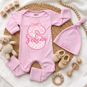 May include: A light pink ribbed long-sleeve romper with a large letter S featuring a floral pattern and a bow, personalised with the name Selena. Includes a matching pink ribbed beanie with the name Selena embroidered in pink script.