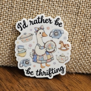 May include: A white die-cut sticker featuring a goose wearing a patchwork dress and a bow, holding an embroidery hoop. The text reads I'd rather be thrifting. Surrounding the goose are illustrations of a casserole dish, a rotary phone, a stack of teacups, a rocking horse, and a teapot.